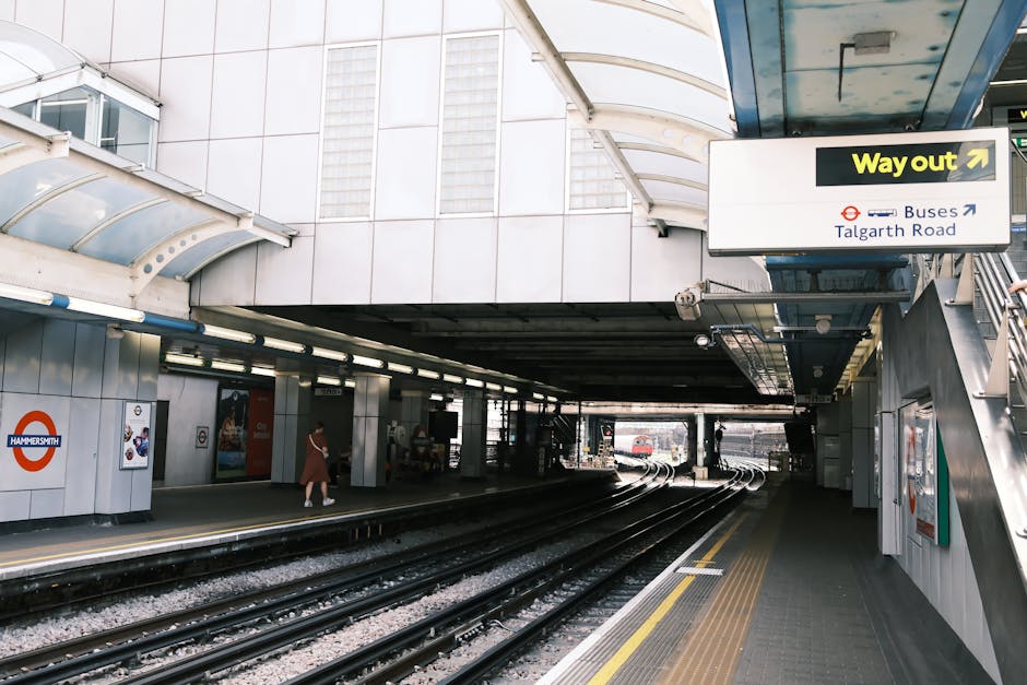 Inside Shoreditch High Street station, a London Underground platform features a long, tiled walkway illuminated by overhead lighting. On the left, a train with yellow and white exterior markings is st